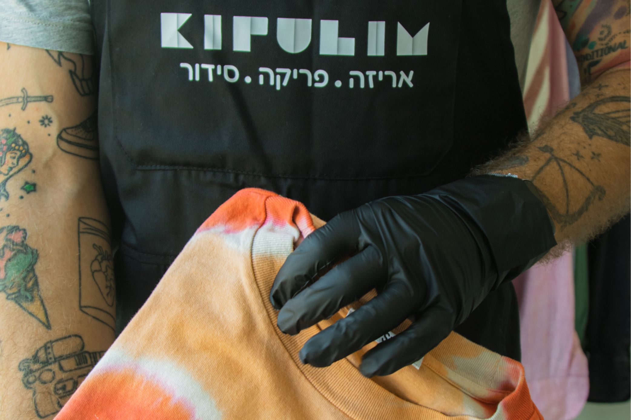 Person wearing a black glove holding an orange tie-dye shirt with 'Kipulim' text on a black background.
